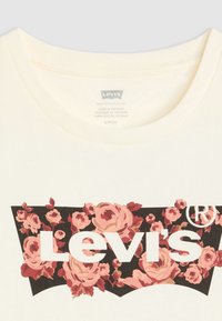 T-shirt de couleur crème avec un graphique noir orné de roses roses et du texte "Levi's" en blanc. Encolure ronde et texture en coton doux.
