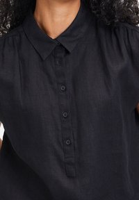 Camicia di lino nera con colletto, cinque bottoni frontali e tessuto morbido con maniche corte. Nessun motivo o accento visibile.