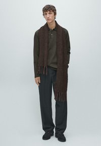 Młody mężczyzna w ciemnooliwkowej koszulce polo, luźnych czarnych spodniach, czarnych butach i długim brązowym szalu stojący na tle jasnego, gładkiego tła.