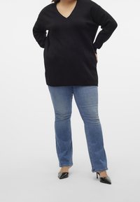Kobieta ubrana w czarny sweter z dekoltem w serek, jasnoniebieskie dżinsy i czarne szpilki, stoi z rękami na biodrach.