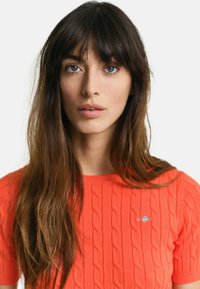 Kortærmet sweater i lys orange med kabelstrik tekstur og et lille broderet logo på brystet. Glat stof med ribbet kant.