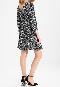 Robe noire à motifs floraux avec des manches trois-quarts, un ourlet évasé et un dos en V. Associée à des talons noirs avec des blocs à pois.