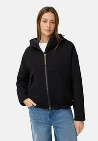Zwarte zip-up hoodie met een gevoerde capuchon, voorzakken en een relaxte pasvorm, gecombineerd met lichtblauwe jeans. Het materiaal lijkt zacht en warm te zijn.