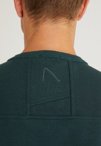 Pohled zezadu na osobu v tmavě zeleném crewnecku s viditelným švem a malým vyšitým trojúhelníkovým logem na horní části zad.