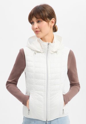 Vrouw draagt een witte doorgestikte mouwloze capuchonvest over een bruin shirt met lange mouwen, handen in de zakken van de vest, kijkt naar de zijkant.
