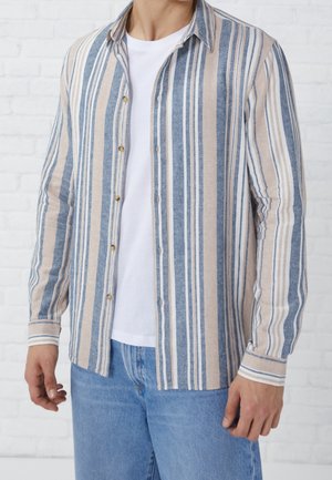 Mand iført beige, blå og hvid vertikalt stribet skjorte over en hvid t-shirt og blå jeans, står mod en hvid murstensvæg.