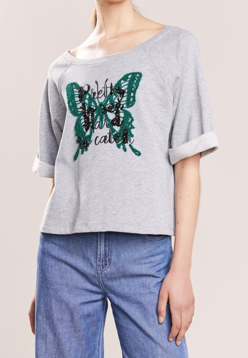 Sweat-shirt gris à manches courtes avec une coupe oversize, arborant un motif de papillon vert et noir avec des sequins et un texte brodé.