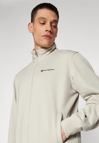 Ljusgrå zip-up jacka med hög krage, med en liten svart och röd Champion-logga på bröstet och sidofickor.
