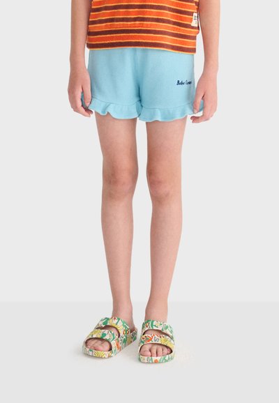 Enfant portant une chemise rayée orange, un short bleu clair à volants et des sandales à enfiler fleuries, debout devant un fond uni.