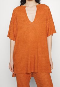 Nainen, jolla on yllään löysä, ribbattu oranssi toppi V-kaula-aukolla ja siihen sopivat ribbatut oranssit housut, näkyy olkapäistä puoliväliin reisiä.