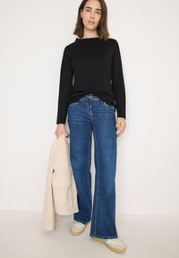 Black long-sleeve top with a round neckline, paired with blue wide-leg jeans. The person holds a beige jacket and wears white sneakers.
