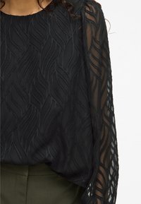 Black blouse featuring a textured wave pattern, sheer long sleeves, and a relaxed fit. Fabric is soft with visible stitching accents and layering.