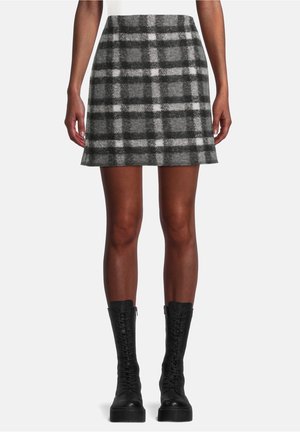 Kvinde iført en grå og hvid ternet mini-nederdel med forreste syninger og sorte snørebånds combat-støvler, stående mod en ensfarvet baggrund.