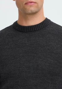 Close-up van de onderkant van het gezicht en de nek van een man die een donkergrijze geribbelde ronde hals trui draagt, op een lichte achtergrond.