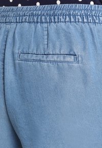 Pantalones de mezclilla azul claro con cintura elástica y un solo bolsillo trasero con detalle de borde cosido. Textura suave.