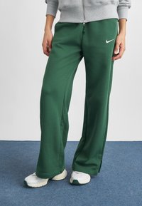 Osoba v zelených teplákoch Nike, sivom zipsovom mikine a bielych teniskách stojí na modrom koberci.