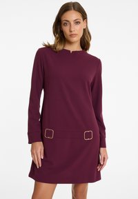 Woman wearing a long-sleeve plum-colored dress with a small front notch neckline and two gold rectangular buckles at the waist.