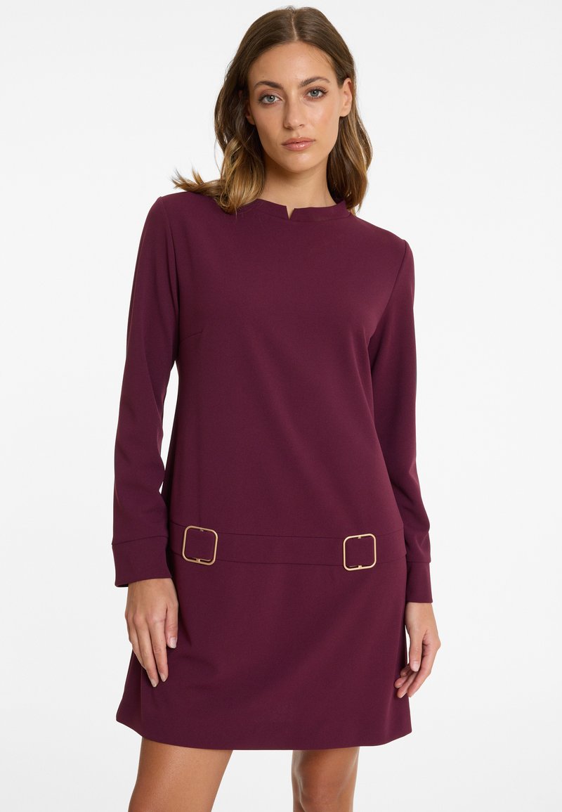 Woman wearing a long-sleeve plum-colored dress with a small front notch neckline and two gold rectangular buckles at the waist.