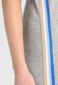 En arm av en person bredvid ett grått tyg med vertikala ränder i beige, vitt, ljusblått och blått.