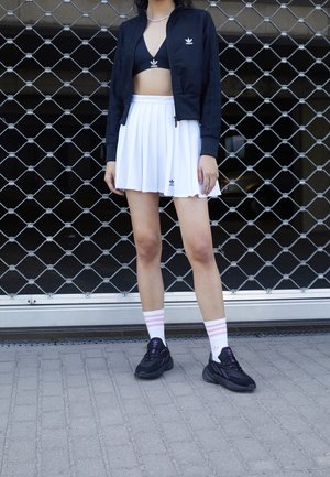 Kobieta ubrana w czarną krótką kurtkę Adidas i sportowy biustonosz, białą plisowaną spódnicę Adidas, białe skarpetki z różowymi paskami oraz czarne trampki, stojąca na zewnątrz.