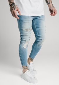 Jeans de mezclilla de color azul claro con detalles desgastados y cortes en las rodillas, combinados con zapatillas blancas. El tejido tiene una textura suave.