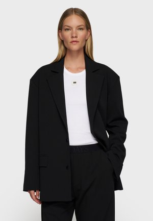 Blondynka w oversize'owym czarnym marynarce, białej dopasowanej koszulce z kwadratową przypinką i czarnych spodniach, stojąca na jasnym, jednolitym tle.