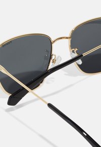 Ochelari de soare cu ramă din metal aurit, lentile gri închise și brațe dinacetat negru. Prezintă suporturi pentru nas clare și un design elegant, unghiular.