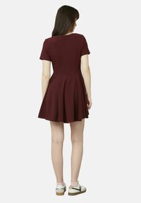 Robe bordeaux à manches courtes avec une jupe évasée, présentant un dos en V et un design plissé. Associée à des chaussures blanches et des chaussettes roses.