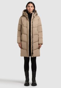 Sieviete, ģērbusies garā bežā pufīgā ziemas mētelī ar kapuci, melnās legingos un melnās zābakos, stāvot uz viendabīgas gaišas fona.