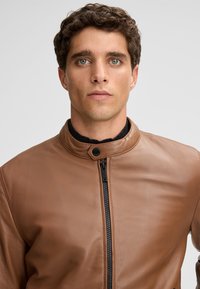 Bruine leren jas met een staande kraag, ritssluiting aan de voorkant en zwarte hardware. Soepele textuur en getailleerde pasvorm.