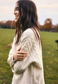 Krém színű kötött pulóver fonott mintával, laza szabású, hosszú ujjú, bordázott mandzsettákkal; fehér felső fölött viselve.