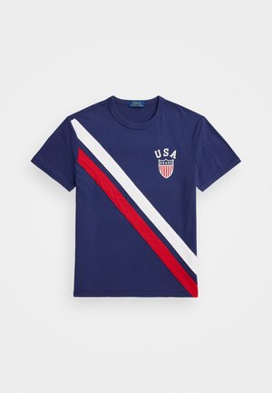 Camiseta navy con rayas rojas y blancas diagonales. Presenta el texto "USA" y un emblema en el pecho. Hecha de algodón, de manga corta y cuello redondo.
