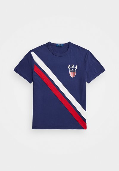 Mornarska majica s diagonalnimi rdečimi in belimi črtami. Na prsih je napis "USA" in grb. Narejena je iz bombaža, s kratkimi rokavi in okroglo ovratnikom.