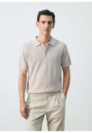 Homme aux cheveux courts et foncés portant un polo en maille beige clair à manches courtes et un pantalon plissé beige clair sur un fond uni.