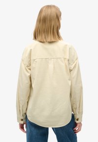 Chemise button-up jaune clair à manches longues, texture à côtes verticales et ourlet arrondi, associée à un jean en denim bleu.