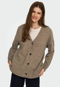 Beige Strickjacke mit V-Ausschnitt, langen Ärmeln, schwarzen Knöpfen und geripptem Saum. Über einem weißen Shirt getragen, mit einer entspannten Passform.