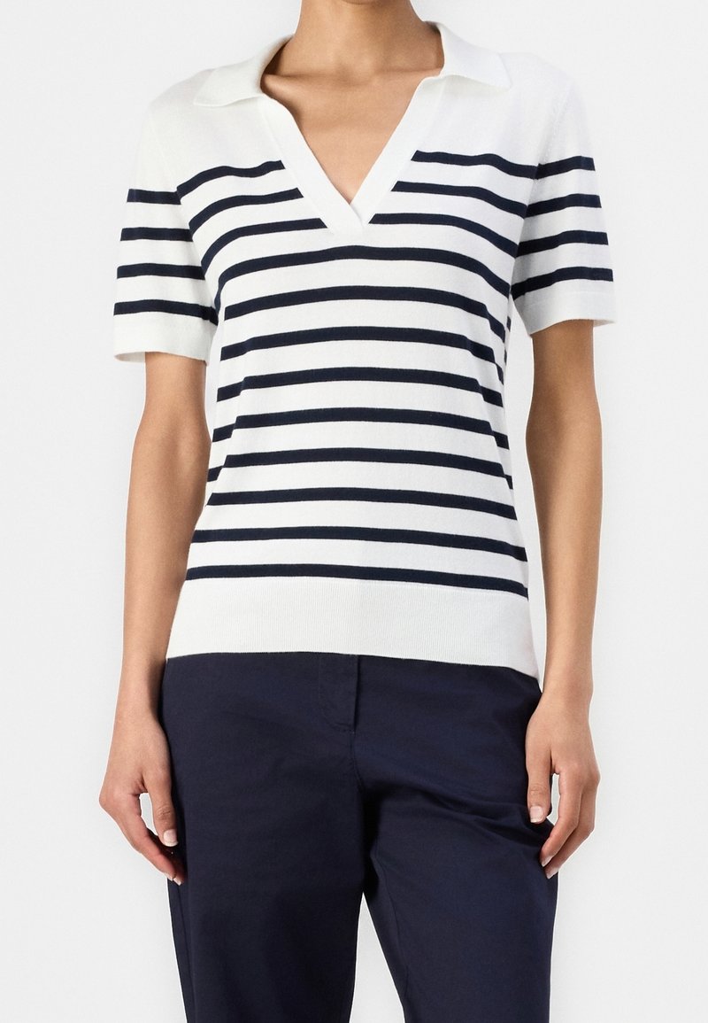 Haut blanc à manches courtes et col en V avec des rayures horizontales bleu marine, porté avec un pantalon bleu marine foncé, visible des épaules jusqu'aux hanches.