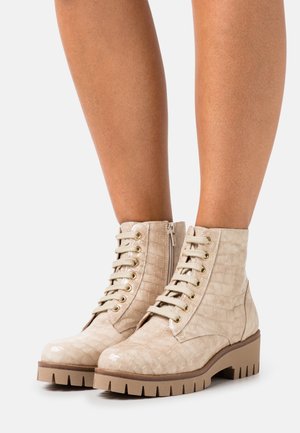 Bottes beiges montantes à la cheville avec motif crocodile, lacets à l'avant, fermeture éclair latérale et semelle épaisse rainurée portées sur des jambes nues.