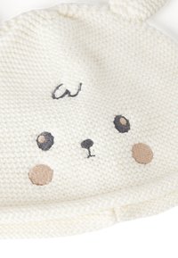 Cappello per bambino lavorato a maglia di colore bianco con design a faccia di animale, occhi e dettagli ricamati. Texture morbida con forma arrotondata e orecchie.