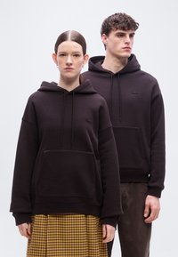 Δύο μοντέλα στέκονται φορώντας ταυτόχρονες σκούρες καφέ φούτερ με κουκούλα, το ένα συνδυασμένο με κίτρινη καρό φούστα, και τα δύο κοιτάζουν προς τα εμπρός με ουδέτερες εκφράσεις.