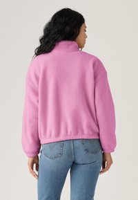 Pull-over en polaire rose avec col haut et coupe décontractée, doté de poignets côtelés et d'un ourlet raccourci, associé à un jean en denim bleu.