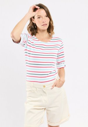 Femme aux cheveux bruns mi-longs portant une chemise blanche avec des rayures rouges, vertes et bleues et un short beige clair, une main dans la poche.