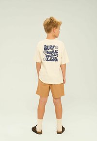 Krämfärgad t-shirt med kort ärm och marinblå grafisk text "SURF MORE WORRY LESS," matchad med bruna shorts och bruna slip-on skor.