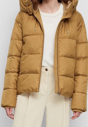 Frau trägt eine braune Steppjacke mit Frontreißverschluss, kombiniert mit hoch taillierten, cremefarbenen Cordhosen und einem weißen Hemd.