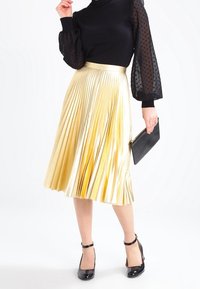 Mujer vistiendo una blusa negra transparente de mangas abullonadas, una falda midi plisada metálica dorada, tacones de charol negros y sosteniendo un bolso de mano negro.