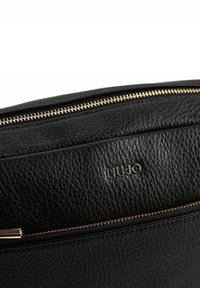 Schwarze Lederhandtasche mit strukturierter Oberfläche, ausgestattet mit einem goldfarbenen Reißverschluss und einem metallischen, auf der Vorderseite geprägten Logo. Kompaktes und schlankes Design.