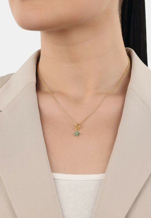 Collar de oro con una cadena delicada y un colgante de piedra preciosa verde con un diseño floral. Lleva puesto sobre un blazer beige y una blusa blanca.