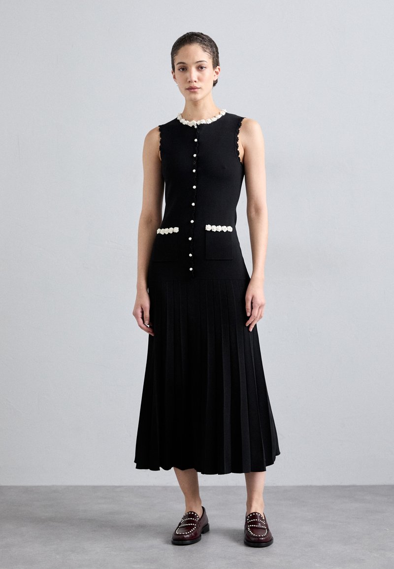 Schwarzes ärmelloses Kleid mit weißem gewelltem Kragen, Perlenknopfdetails und zwei Taschen; hat einen plissierten Rock. Getragen mit dunklen Slippern.