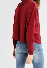 Blusa bordeaux con colletto plissettato, vestibilità ampia, maniche lunghe e dettagli in bottone sui polsini, abbinata a jeans in denim blu aderenti.