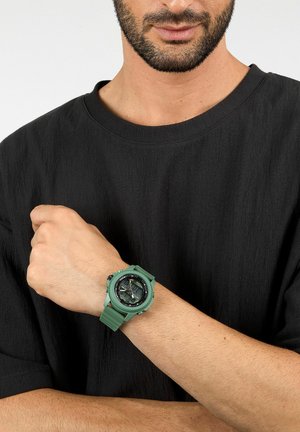 Orologio in gomma verde con quadrante rotondo, display analogico e digitale, cinturino texturizzato e quadrante nero a contrasto, indossato su una mano contro una camicia nera.